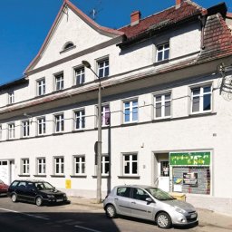 AGWA Sp. z o.o. - Trzypiętrowy budynek z jasną elewacją, widok z ulicy z zaparkowanymi samochodami, widoczne okna z białymi ramami i częściowo odnowiony dach z czerwonej dachówki.