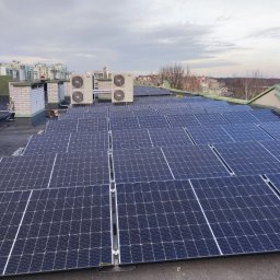 AGWA Sp. z o.o. - Instalacja paneli fotowoltaicznych na płaskim dachu budynku, widoczne klimatyzatory i fragmenty zabudowy miejskiej w tle.