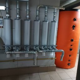 AGWA Sp. z o.o. - Szereg pionowych filtrów z metalowymi rurami i pomarańczowy zasobnik ciepłej wody w pomieszczeniu z kafelkami.