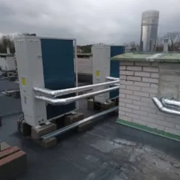 Dwa agregaty klimatyzacyjne zamontowane na płaskim dachu, rury w izolacji termicznej prowadzą do komina z cegły