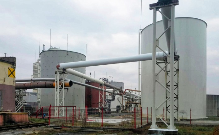 Zewnętrzny widok na przemysłowe zbiorniki cylindryczne połączone rurociągami, z widoczną konstrukcją wsporczą i ogrodzeniem z czerwonej siatki na pierwszym planie.