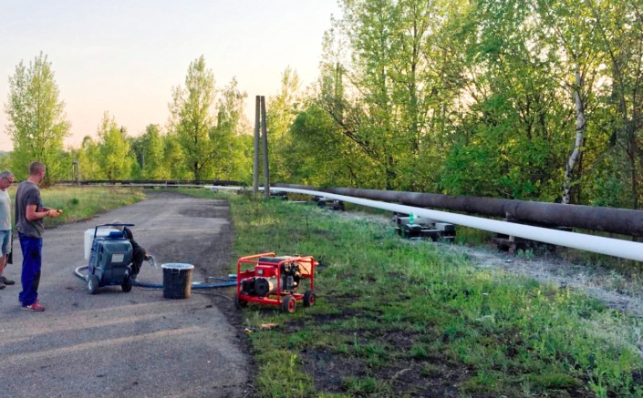 Malowanie rurociągu nadziemnego, widoczny agregat malarski, generator prądu i dwóch mężczyzn podczas pracy.