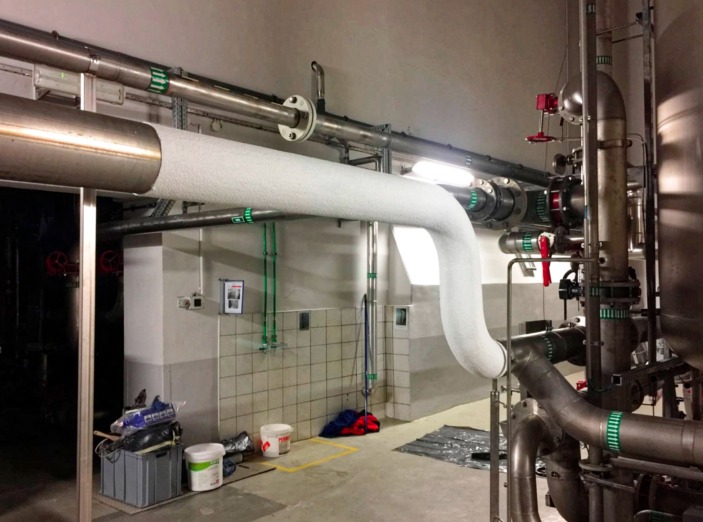 Izolowane rury instalacji grzewczej w kotłowni, widoczne detale łączeń i zaworów, ściany wyłożone częściowo płytkami.