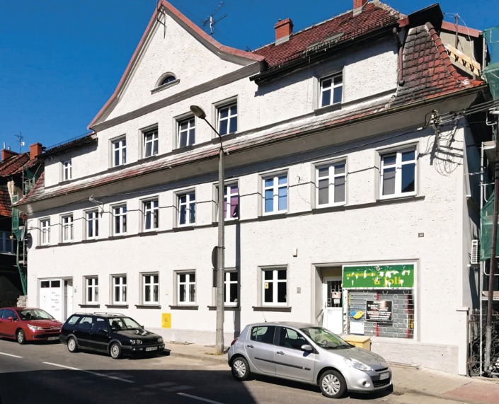 Trzypiętrowy budynek z jasną elewacją, widok z ulicy z zaparkowanymi samochodami, widoczne okna z białymi ramami i częściowo odnowiony dach z czerwonej dachówki.