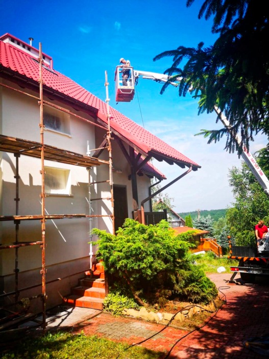 Pracownik na podnośniku koszowym wykonuje prace na elewacji domu z czerwoną dachówką, widoczne rusztowanie i przygotowana ściana, w tle zieleń drzew i błękitne niebo.