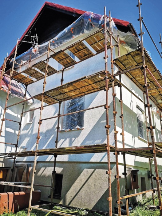 Dom jednorodzinny w trakcie ocieplania, zabezpieczony rusztowaniem i folią ochronną na oknach, z widocznym fragmentem dachu pokrytego czerwoną dachówką.