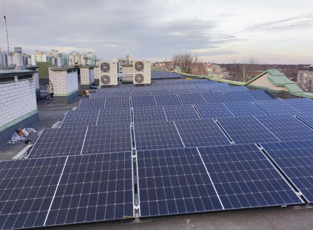 Instalacja paneli fotowoltaicznych na płaskim dachu budynku, widoczne klimatyzatory i fragmenty zabudowy miejskiej w tle.