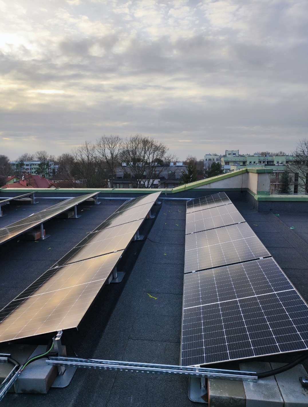 Rząd paneli słonecznych na płaskim dachu pokrytym czarną papą, widok z góry, pochmurne niebo w tle.
