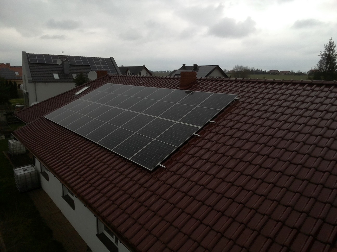 Panele słoneczne zamontowane na brązowym, spadzistym dachu domu jednorodzinnego, widok z góry na tle pochmurnego nieba.