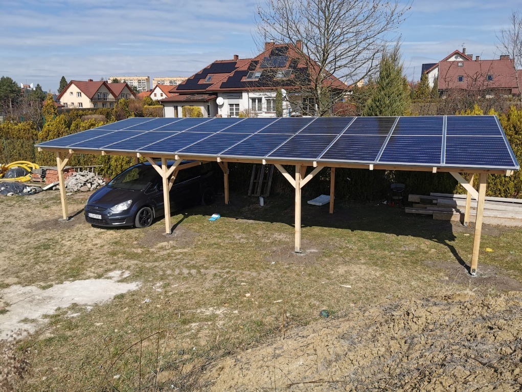 Drewniana wiata garażowa z panelami słonecznymi na dachu, pod którą zaparkowany jest samochód osobowy, na tle domów jednorodzinnych z instalacjami fotowoltaicznymi na dachach.