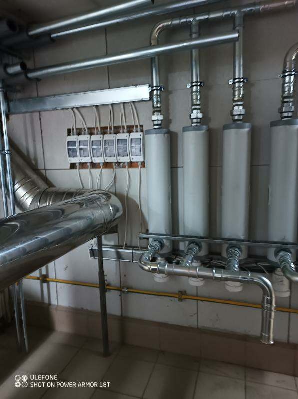 Szereg pionowych filtrów cylindrycznych z metalowymi rurami i elementami mocującymi, widoczne także przewody elektryczne i rura wentylacyjna na tle ściany z ceramicznych płytek.