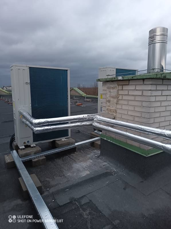 Klimatyzatory przemysłowe na dachu budynku z izolowanymi rurami, widok na komin i pochmurne niebo.