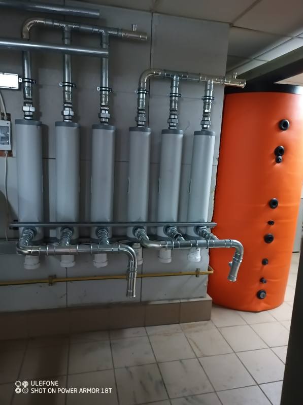 Szereg pionowych filtrów z metalowymi rurami i pomarańczowy zasobnik ciepłej wody w pomieszczeniu z kafelkami.