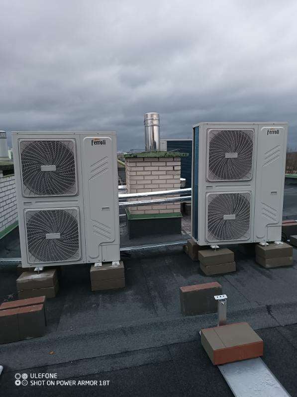 Dwa klimatyzatory Ferroli zainstalowane na dachu budynku w Warszawie, umieszczone na ceglanych podstawach, z kominem w tle i pochmurnym niebem.