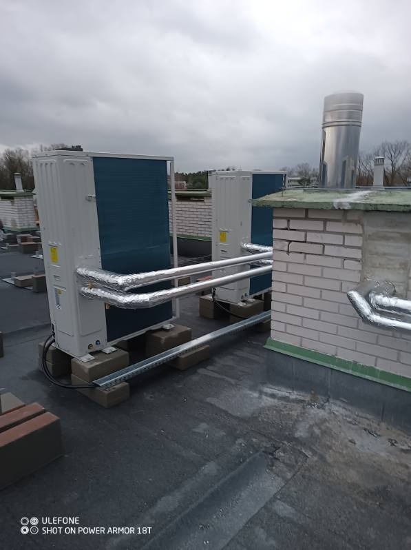 Dwa agregaty klimatyzacyjne zamontowane na płaskim dachu, rury w izolacji termicznej prowadzą do komina z cegły