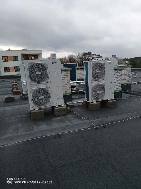 Dwa klimatyzatory marki Ferroli zamontowane na płaskim dachu budynku, każdy na podstawie z cegieł, widoczne przewody i fragmenty innych urządzeń w tle, pochmurne niebo.