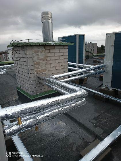 Instalacja grzewcza na dachu budynku z kominem, rury w srebrnej izolacji termicznej, widoczne jednostki zewnętrzne klimatyzacji.