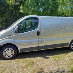 Srebrny bus Renault Trafic zaparkowany na trawiastym podłożu, widok z boku na tle zieleni drzew.