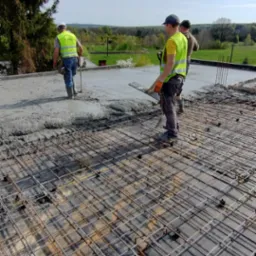 Trzech robotników w kamizelkach odblaskowych rozprowadza mokry beton na siatce zbrojeniowej na dachu budynku, widok na zielone wzgórza w tle.