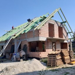 R-PRO KAROLINA RURARZ - Budowa domu jednorodzinnego w trakcie realizacji: ściany z czerwonej cegły, widoczne rusztowania, mieszalnik do betonu, palety, pracownik na dachu montujący konstrukcję drewnianą. Na pierwszym...