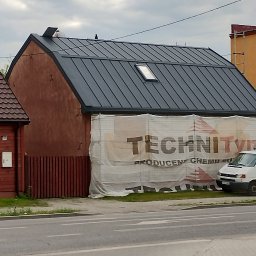 R-PRO KAROLINA RURARZ - Dom z nowym, grafitowym dachem blachodachówkowym i oknem dachowym, częściowo zasłonięty banerem reklamowym. Widok z poziomu ulicy.