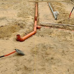 R-PRO KAROLINA RURARZ - Pomarańczowe rury kanalizacyjne ułożone w wykopie pod fundamenty domu. W tle widać pracownika i zaparkowaną ciężarówkę.