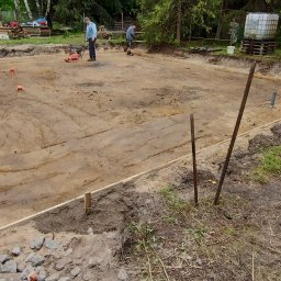 R-PRO KAROLINA RURARZ - Przygotowanie terenu pod fundamenty: ubijanie ziemi zagęszczarką, widoczne drewniane szalunki i wytyczone granice. Dwóch robotników w trakcie pracy.