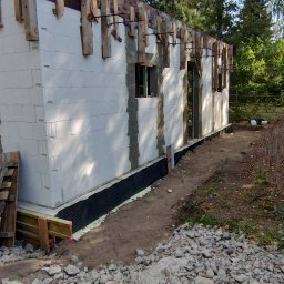 R-PRO KAROLINA RURARZ - Budowa domu z białych bloczków, widoczne drewniane szalunki i izolacja fundamentów. W tle drzewa i niebo. Częściowo ułożone zbrojenie.