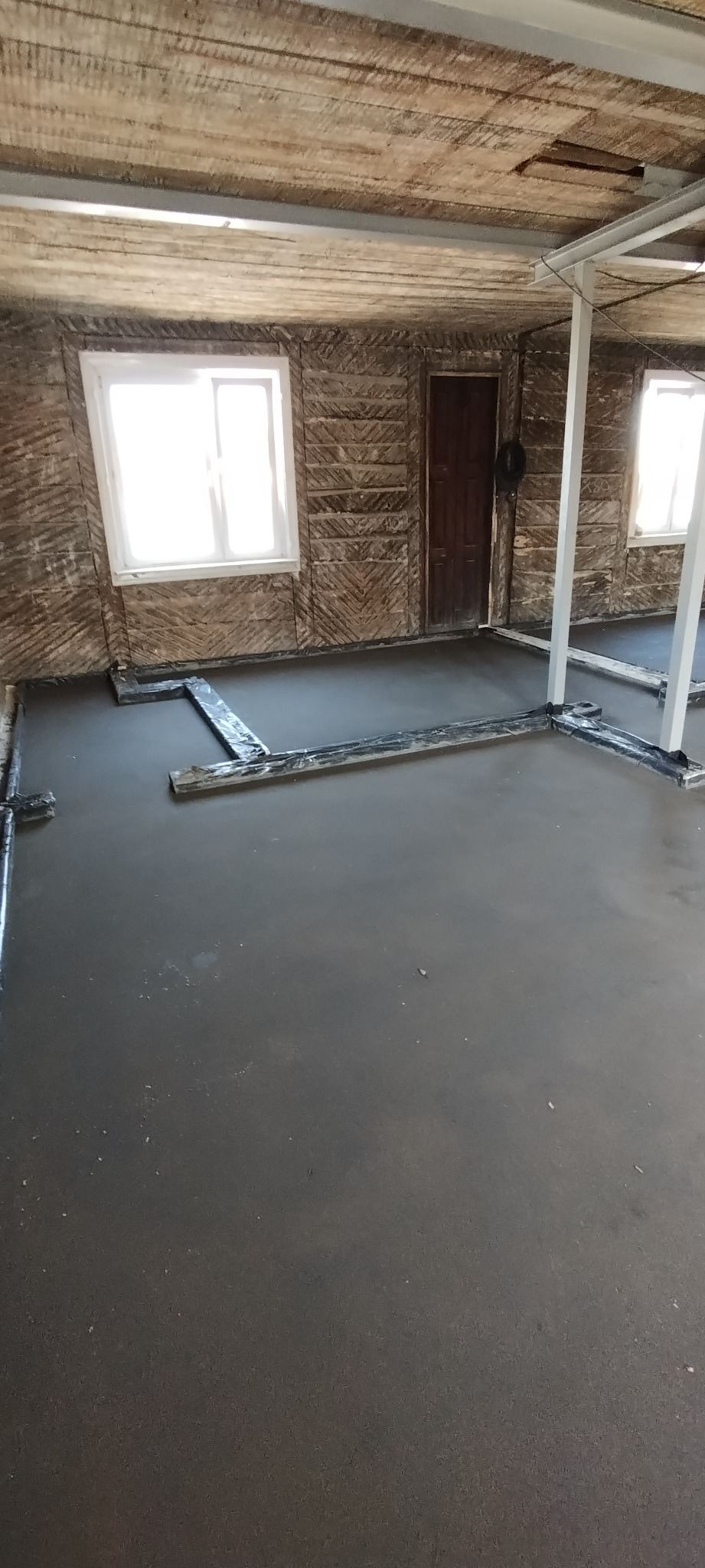 Wylana, świeża wylewka betonowa na podłodze wewnątrz budynku z surowymi ścianami i oknami. Widoczne listwy dylatacyjne i podpory konstrukcyjne.