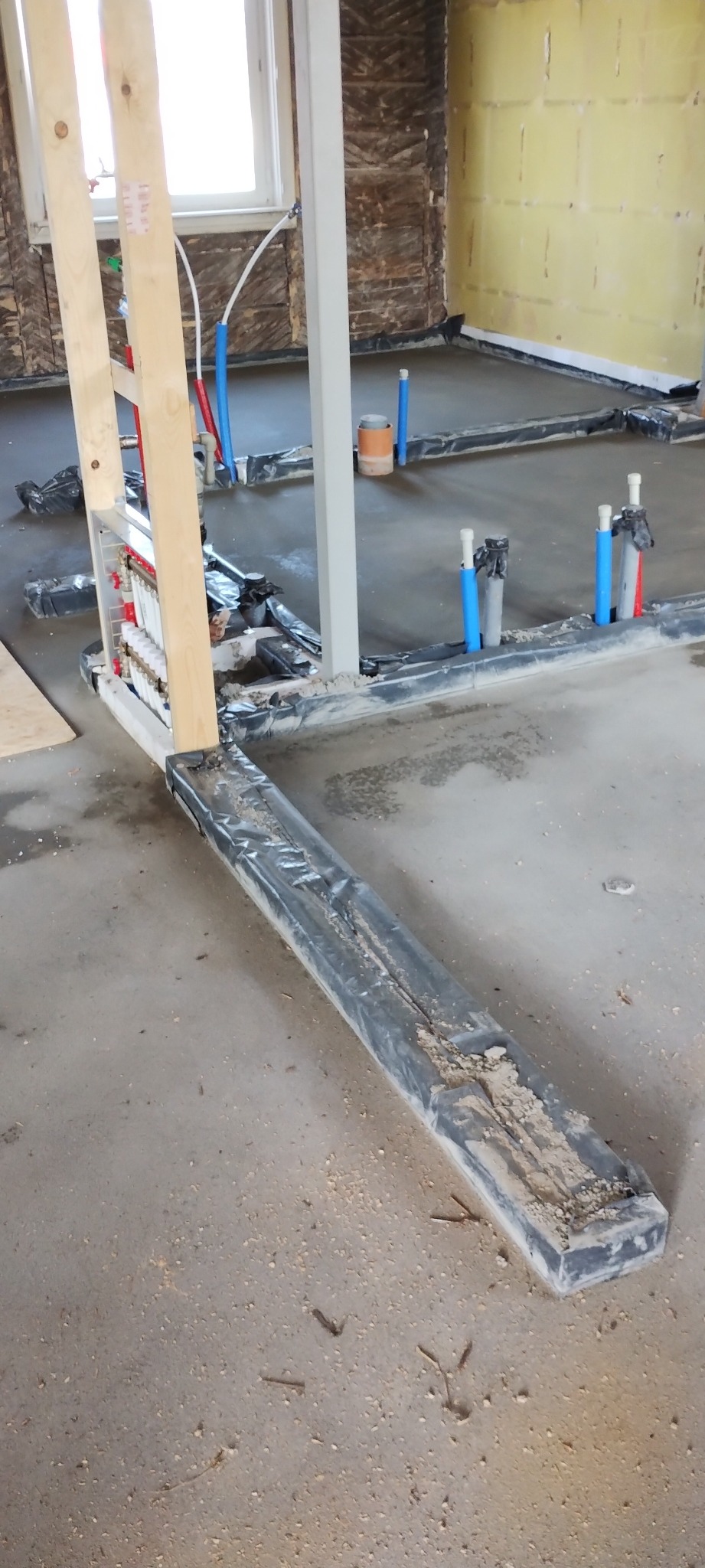 Instalacja podłogowa w trakcie montażu: rury grzewcze, izolacja, rozdzielacz. Widoczne elementy konstrukcyjne ścian i okno. Szary beton na podłodze.