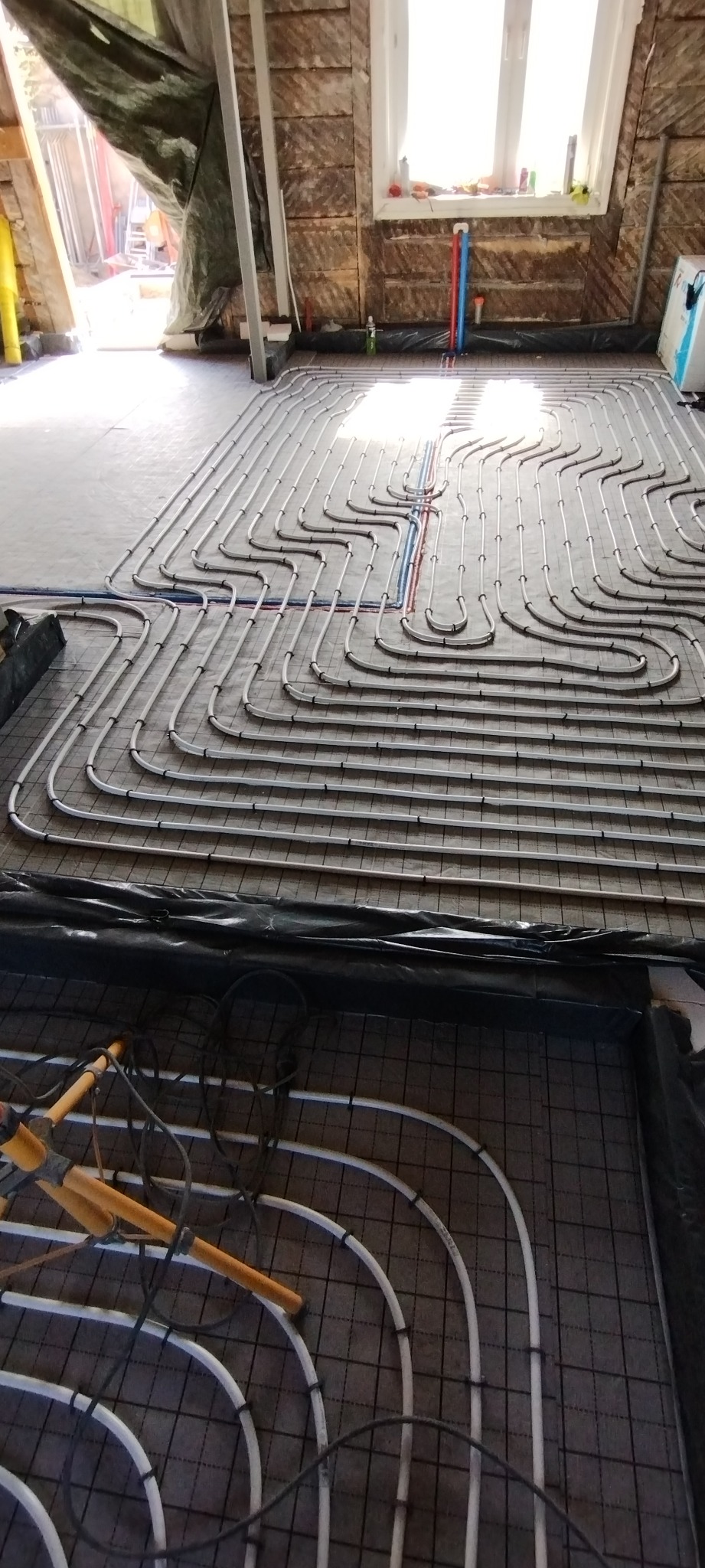 Instalacja ogrzewania podłogowego w poddaszu: białe rury ułożone w pętle na czarnej macie, widoczne narzędzia, okno i ocieplone ściany. Etap prac przed wylewką.