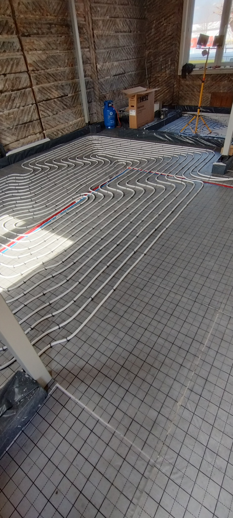 Ułożone rury ogrzewania podłogowego na podkładzie z siatką w nowym budynku. Widoczna instalacja przed wylaniem posadzki, niebieska butla gazowa i lampa budowlana.