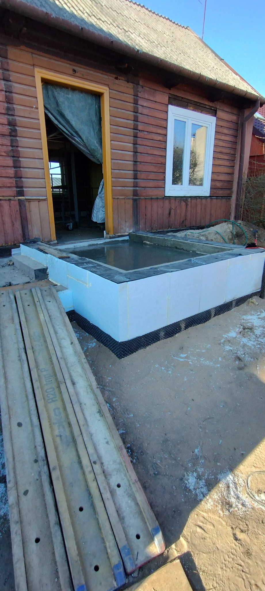 Fundamenty domu z drewnianą elewacją, widoczne szalunki i izolacja termiczna. Wnętrze fundamentu wypełnione wodą, co sugeruje test szczelności.