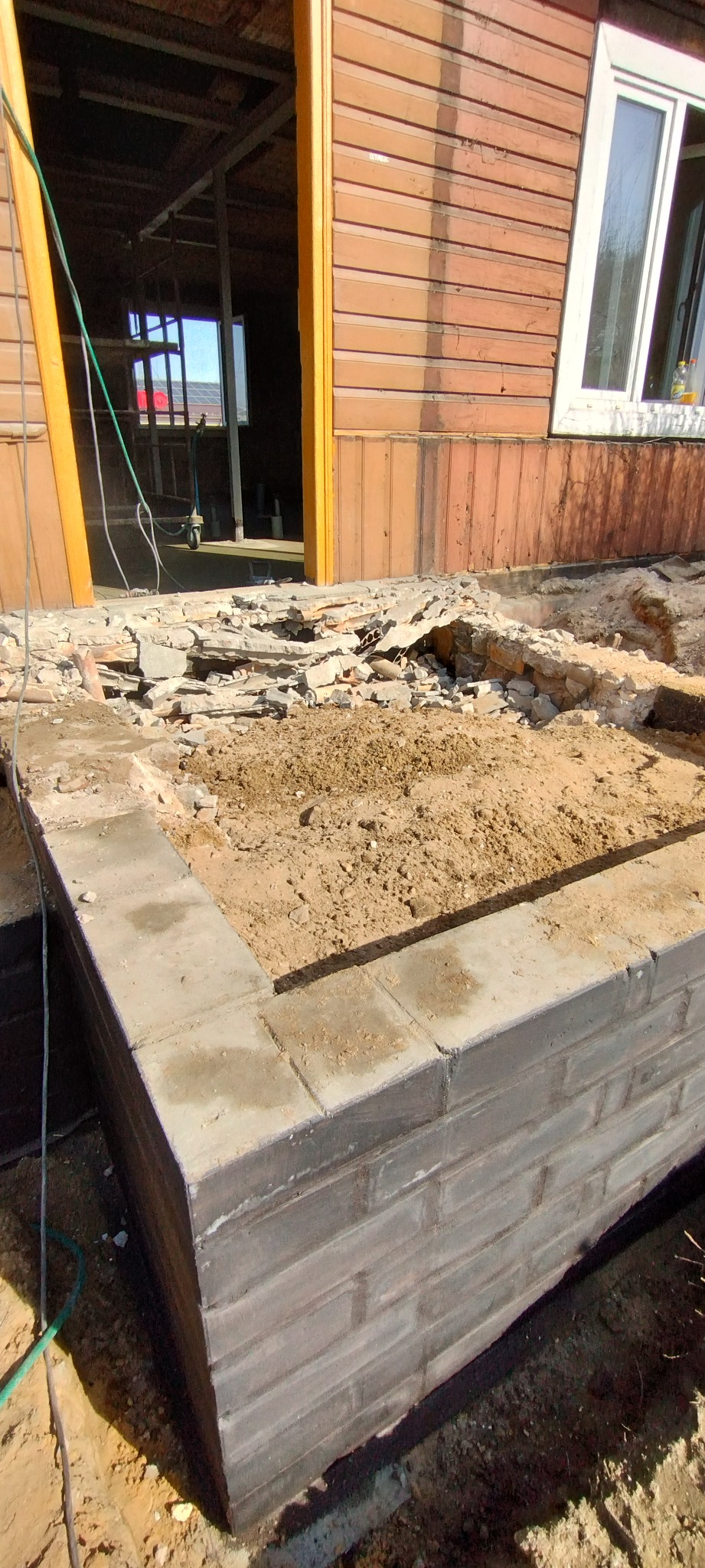 Wznoszenie fundamentu pod budynek z drewnianą elewacją. Widoczne bloczki fundamentowe i piasek. W tle otwór drzwiowy i okno.