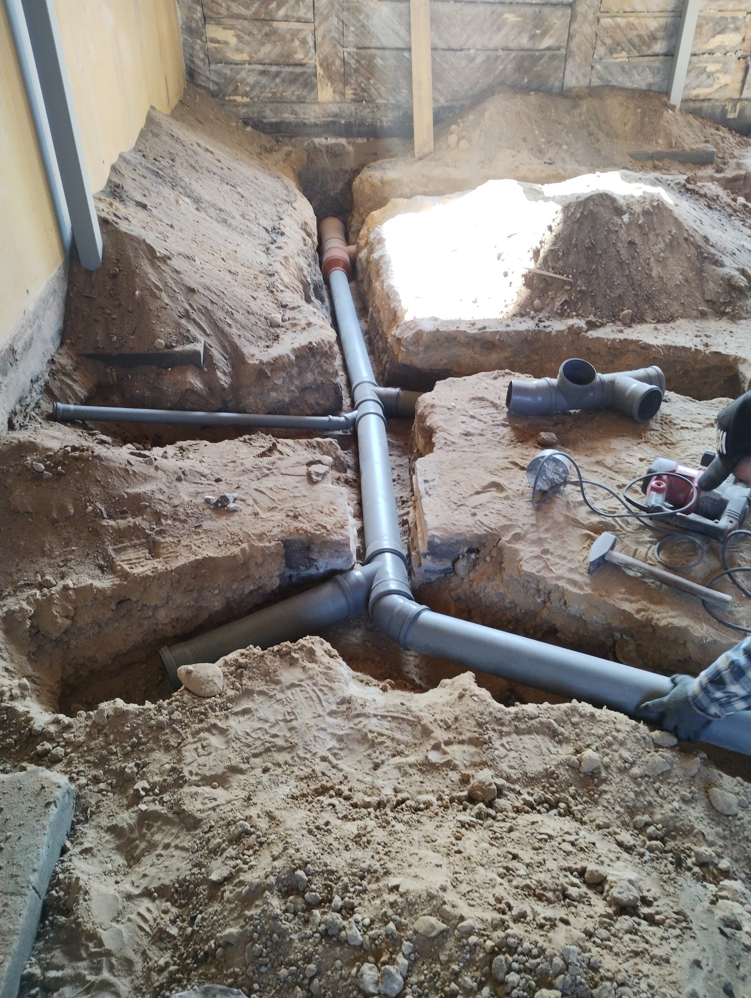 Szare rury kanalizacyjne ułożone w wykopie fundamentowym z piaskiem. Widoczne narzędzia i fragment dłoni w roboczej rękawicy. Etap prac instalacyjnych.