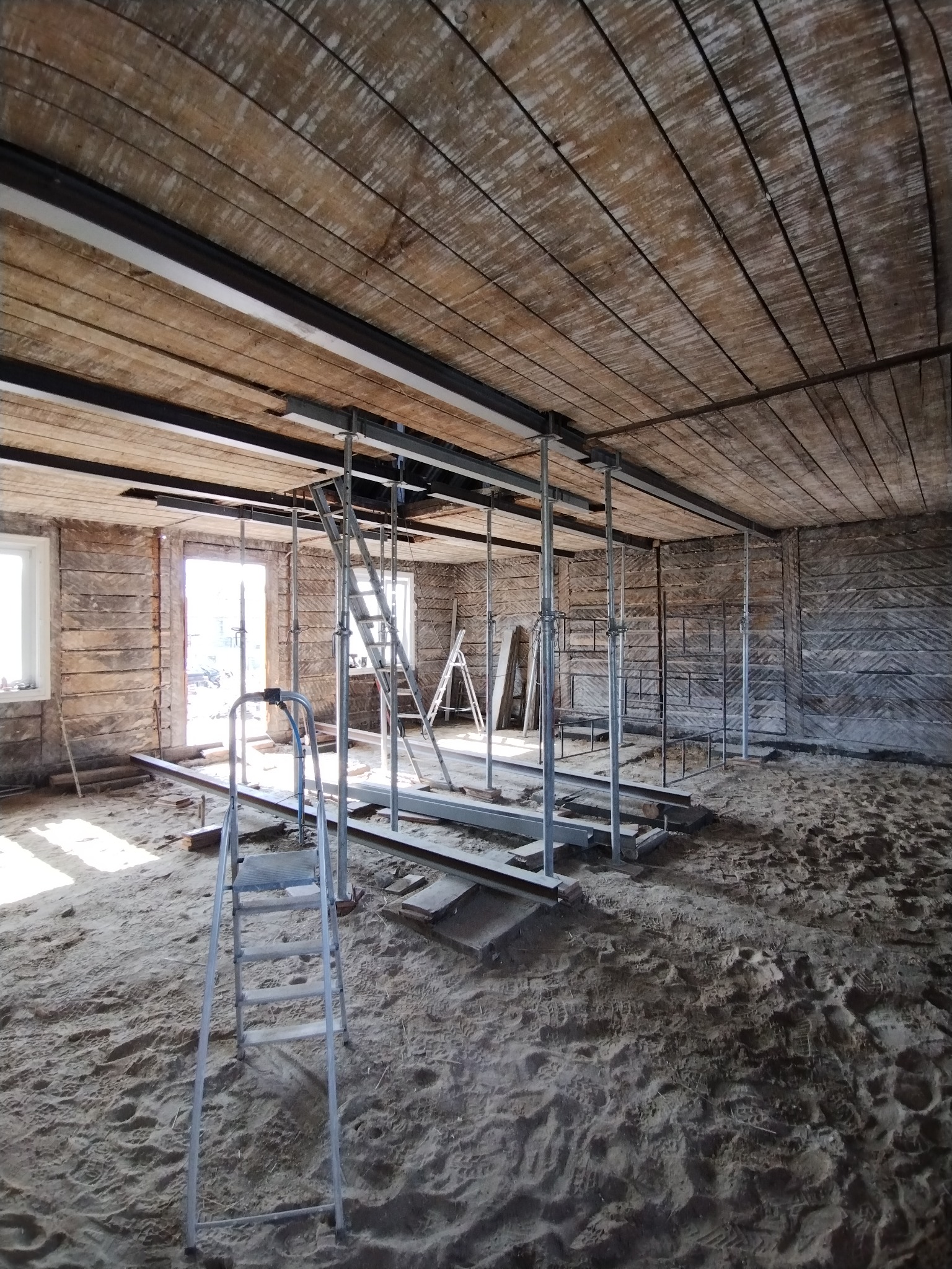 Wnętrze remontowanego domu z drewnianymi ścianami i stropem, podparte metalowymi stemplami. Widoczne drabiny i piasek na podłodze.