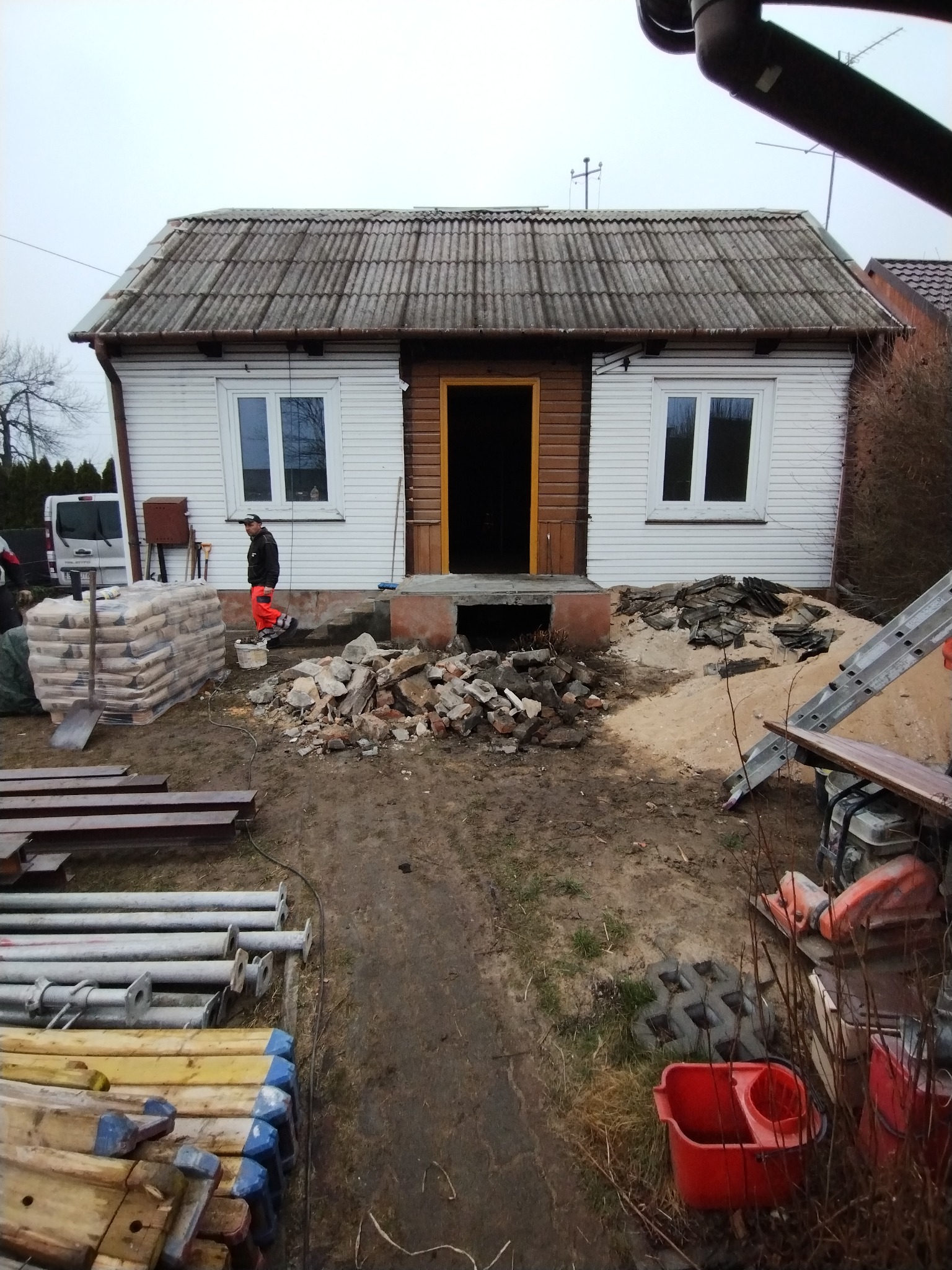 Demontaż starego wejścia do domu z eternitowym dachem. Na pierwszym planie gruz, worki z zaprawą i elementy konstrukcyjne. Robotnik w stroju roboczym w tle.