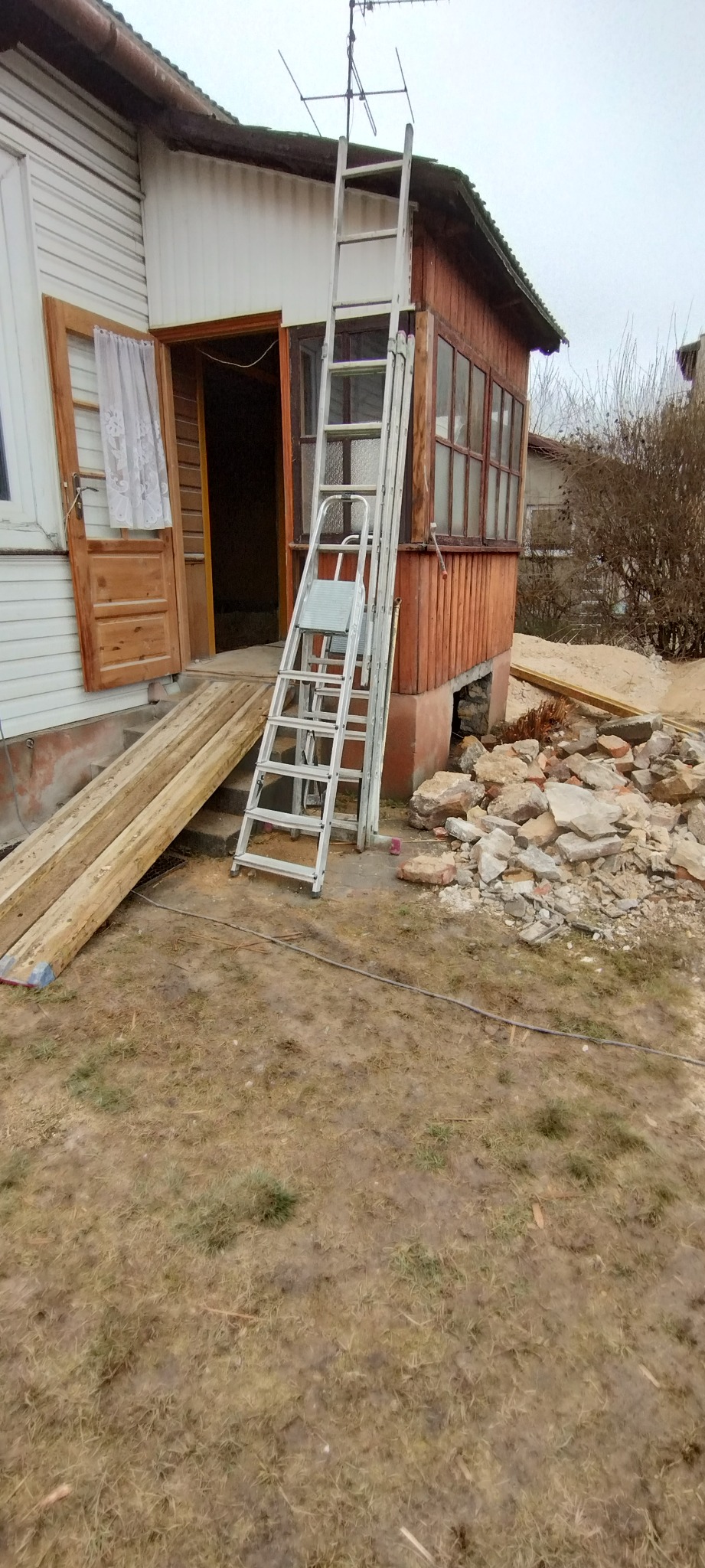 Remont domu: drabina oparta o werandę z oknami, deski i gruz na trawie. Otwarta drewniana brama i okno z firanką.