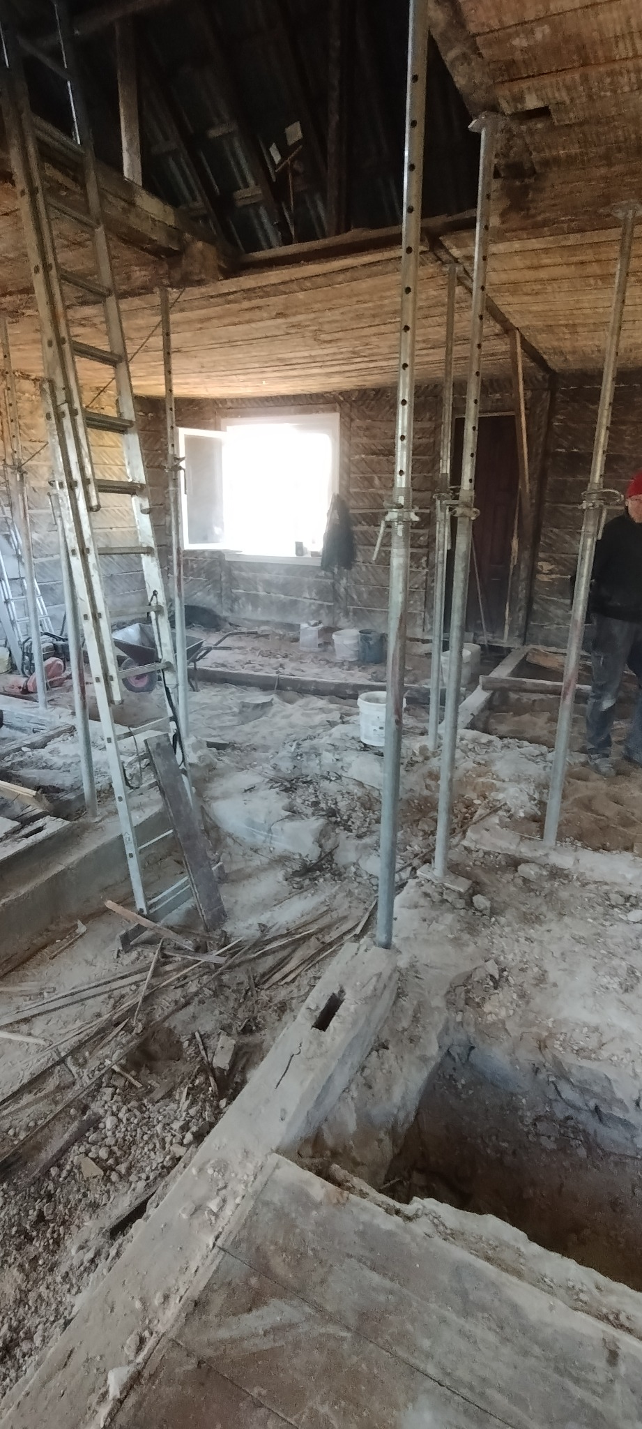 Wnętrze remontowanego poddasza z podporami stropu i drabiną, widoczne narzędzia i materiały budowlane, okno z jasnym światłem.