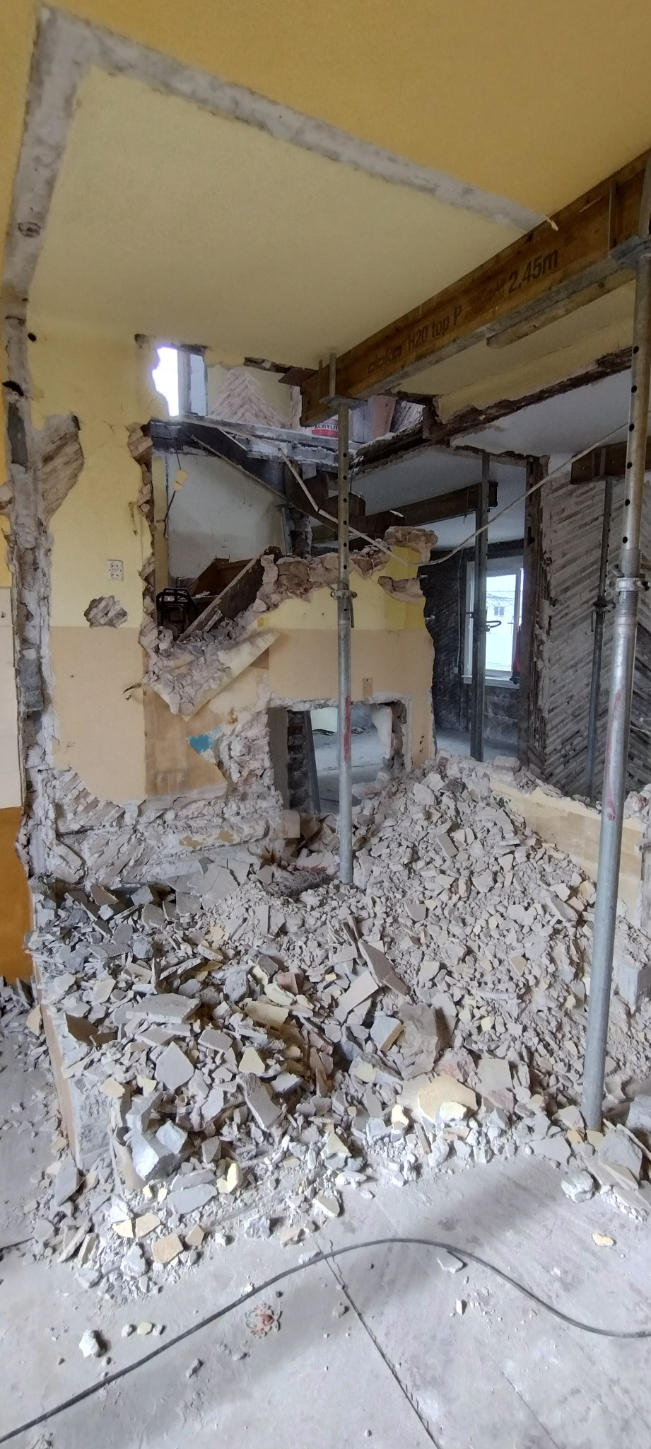 Wnętrze w trakcie rozbiórki ścian, zabezpieczone metalowymi stemplami. Gruz na podłodze, odsłonięte fragmenty instalacji. Widoczne okno w tle.
