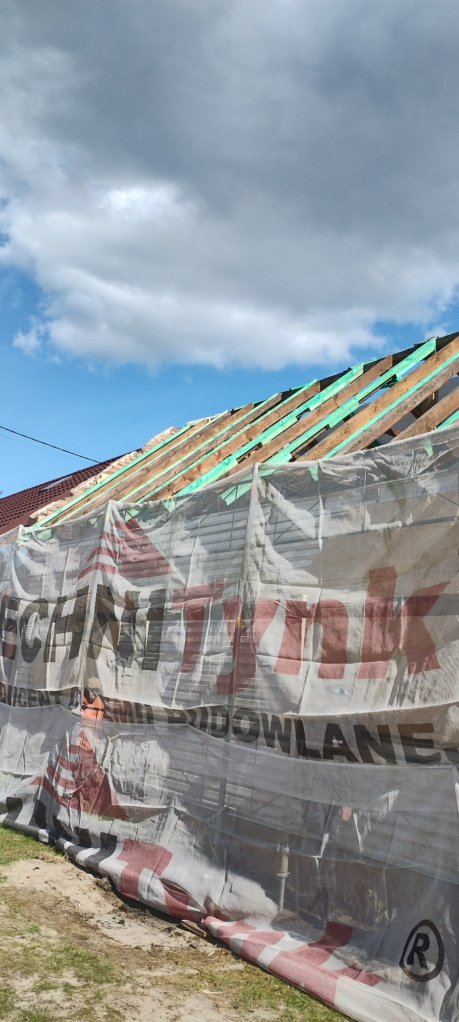 Konstrukcja dachu w trakcie budowy, zabezpieczona siatką ochronną z widocznym logo producenta. Drewniane elementy konstrukcyjne i fragment nieba z chmurami w tle.