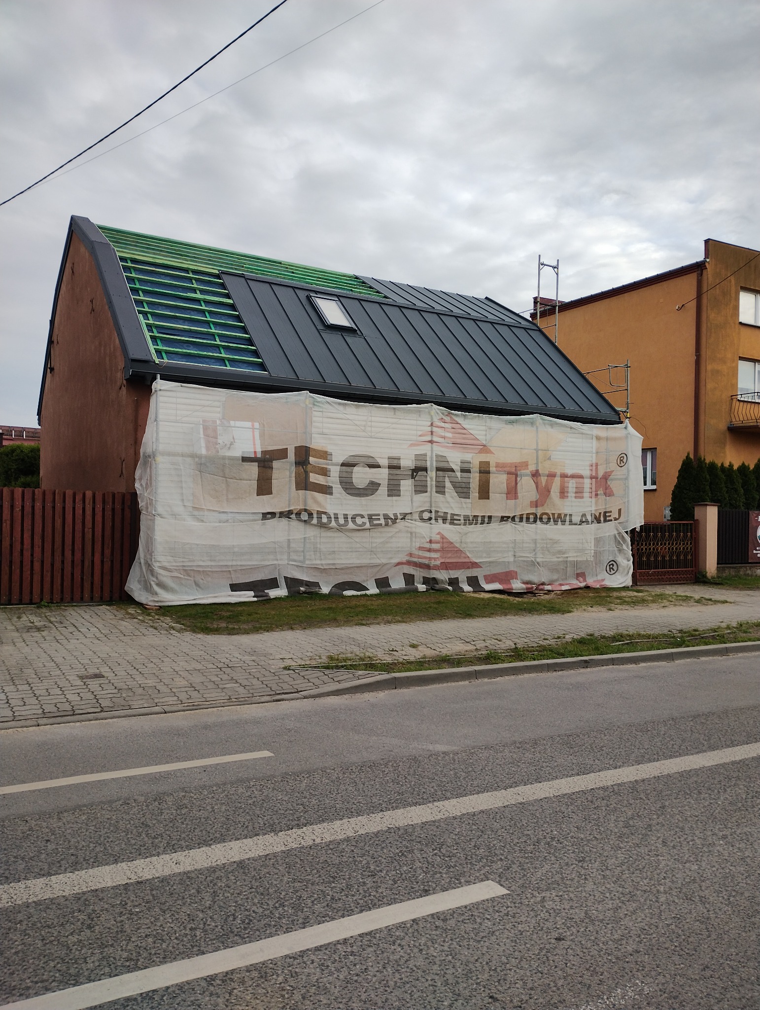 Dom w trakcie budowy dachu, widoczne drewniane elementy konstrukcji i pokrycie blachodachówką, osłonięty banerem reklamowym producenta chemii budowlanej.