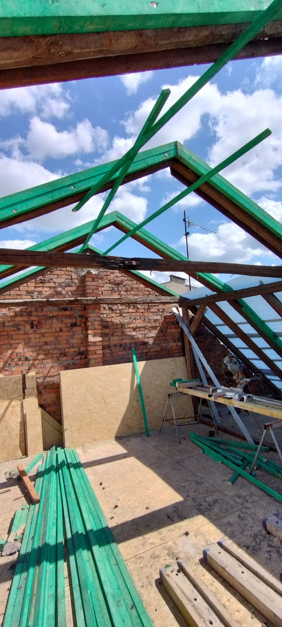 Wnętrze domu w trakcie budowy dachu: widoczne krokwie pomalowane na zielono, ściana z czerwonej cegły, płyty OSB i narzędzia. W tle błękitne niebo.