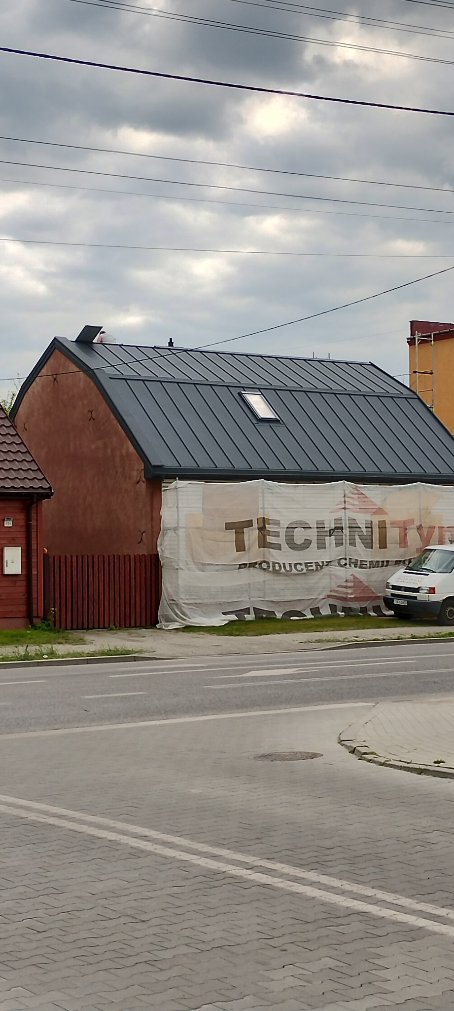 Dom z nowym, grafitowym dachem blachodachówkowym i oknem dachowym, częściowo zasłonięty banerem reklamowym. Widok z poziomu ulicy.