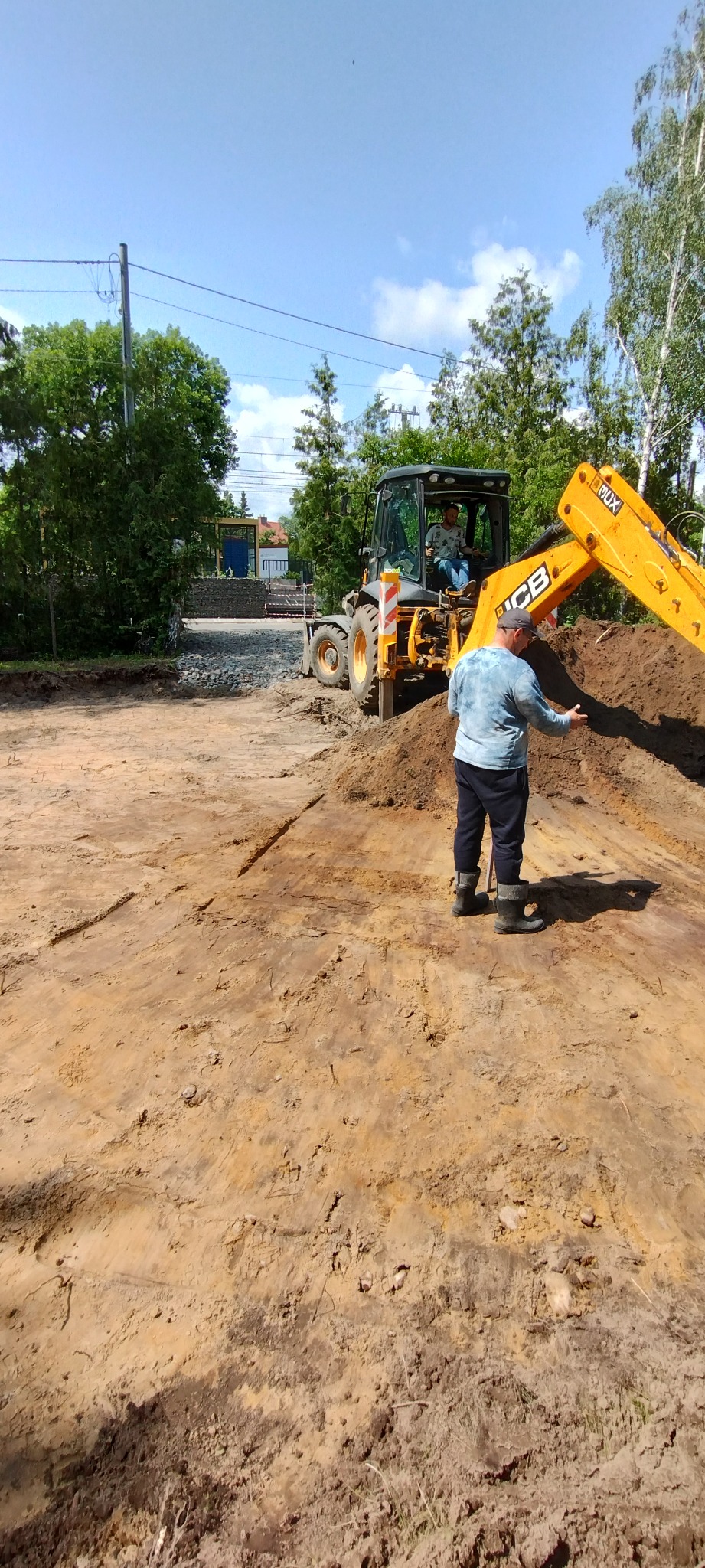 Praca ziemne z użyciem koparko-ładowarki JCB. Operator w kabinie, mężczyzna w roboczych butach i czapce kontroluje prace ziemne, teren wyrównany. Słoneczny dzień.