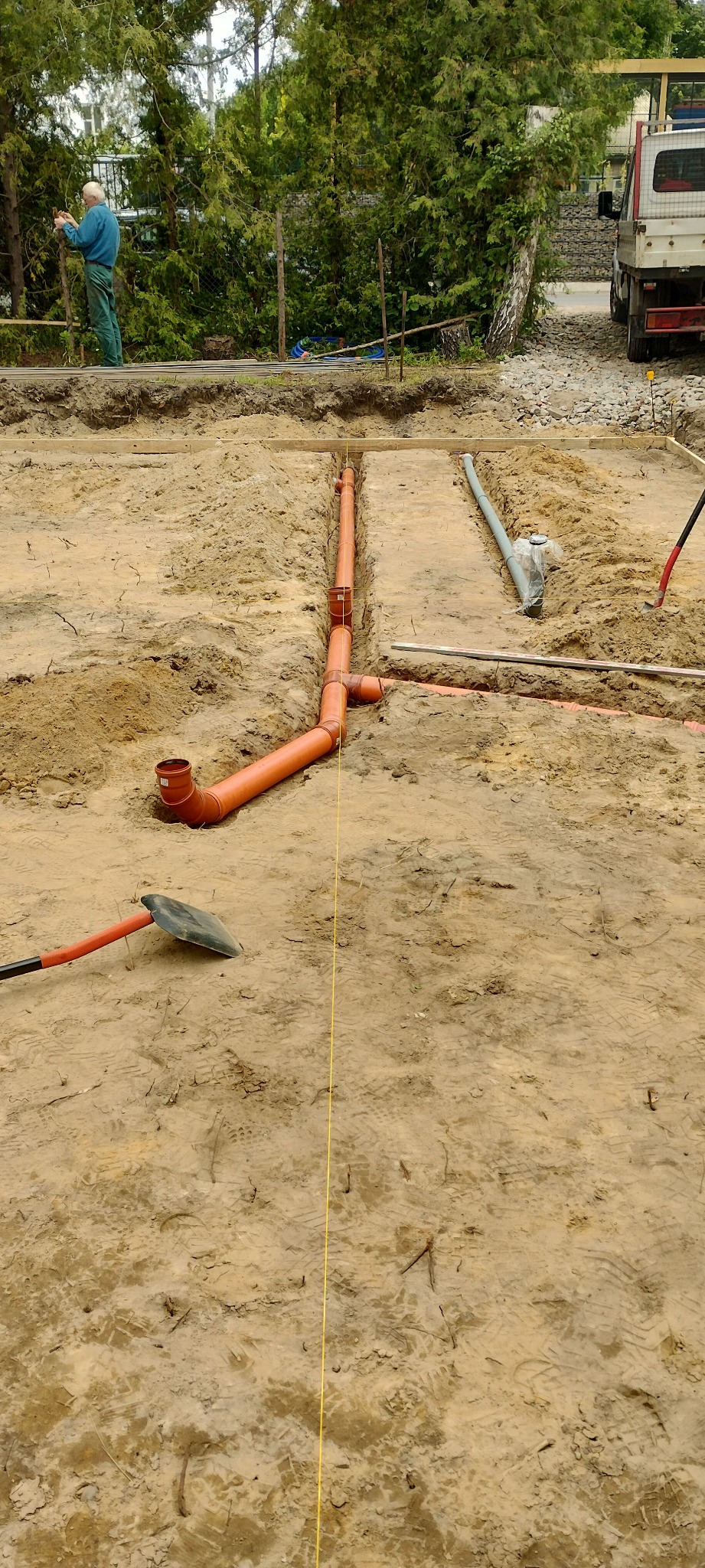 Pomarańczowe rury kanalizacyjne ułożone w wykopie pod fundamenty domu. W tle widać pracownika i zaparkowaną ciężarówkę.
