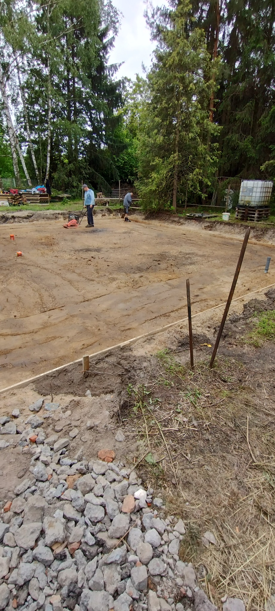 Przygotowanie terenu pod fundamenty: ubijanie ziemi zagęszczarką, widoczne drewniane szalunki i wytyczone granice. Dwóch robotników w trakcie pracy.