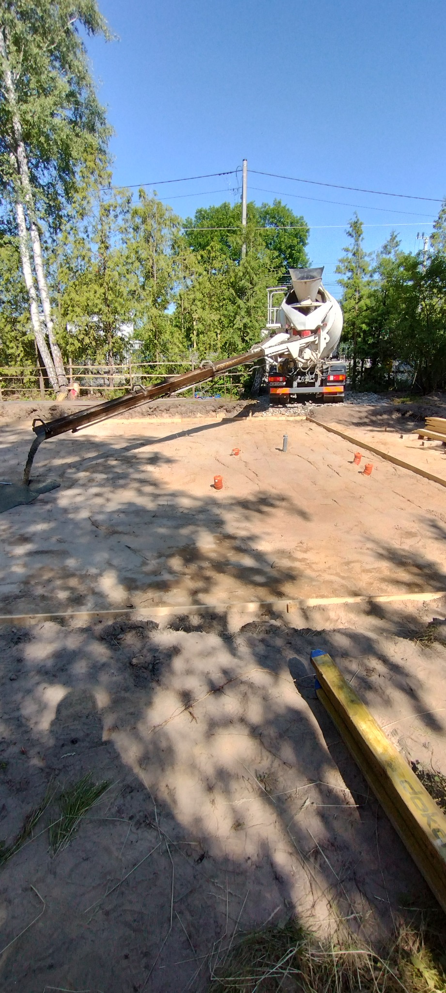 Wylewanie betonu na przygotowany fundament pod dom. Widoczna pompa do betonu i drewniane szalunki. Praca w toku, słoneczny dzień.