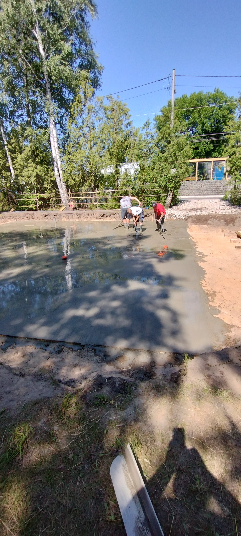 Wylewka fundamentu z pracującymi zbrojarzami. Widoczne narzędzia i teren budowy w słoneczny dzień. Prace ziemne w toku.