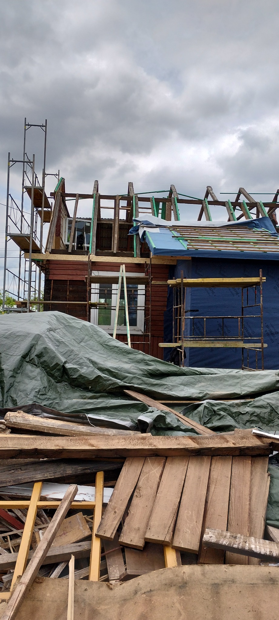 Rozbudowa domu z drewnianą konstrukcją dachu, zabezpieczona folią i rusztowaniami. Na pierwszym planie składowane deski i odpady budowlane.